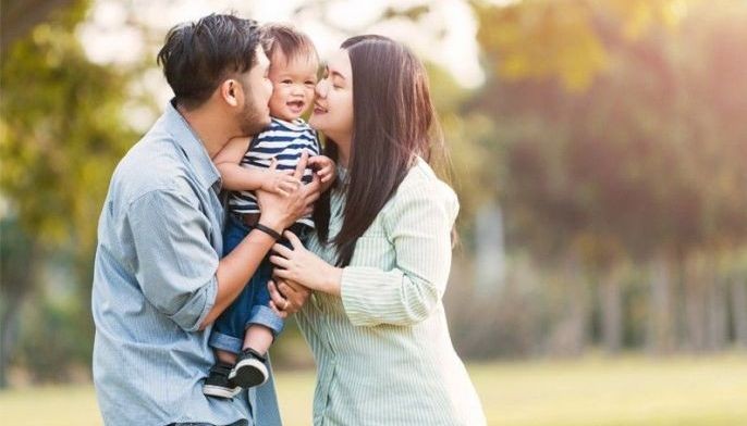 A mother and father kissing their smiling baby in a sunny park